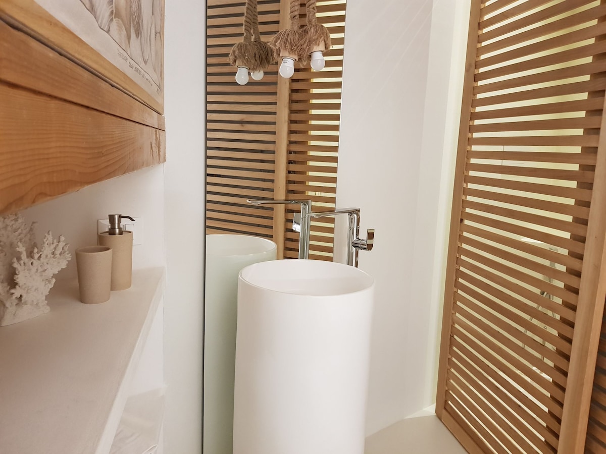A modern bathroom is highlighted by a freestanding round sink, complemented by wooden features and vertical slatted panels. Soft lighting enhances the neutral color palette, creating a calm ambiance. A large mirror reflects the space, adding depth and light to the area.
