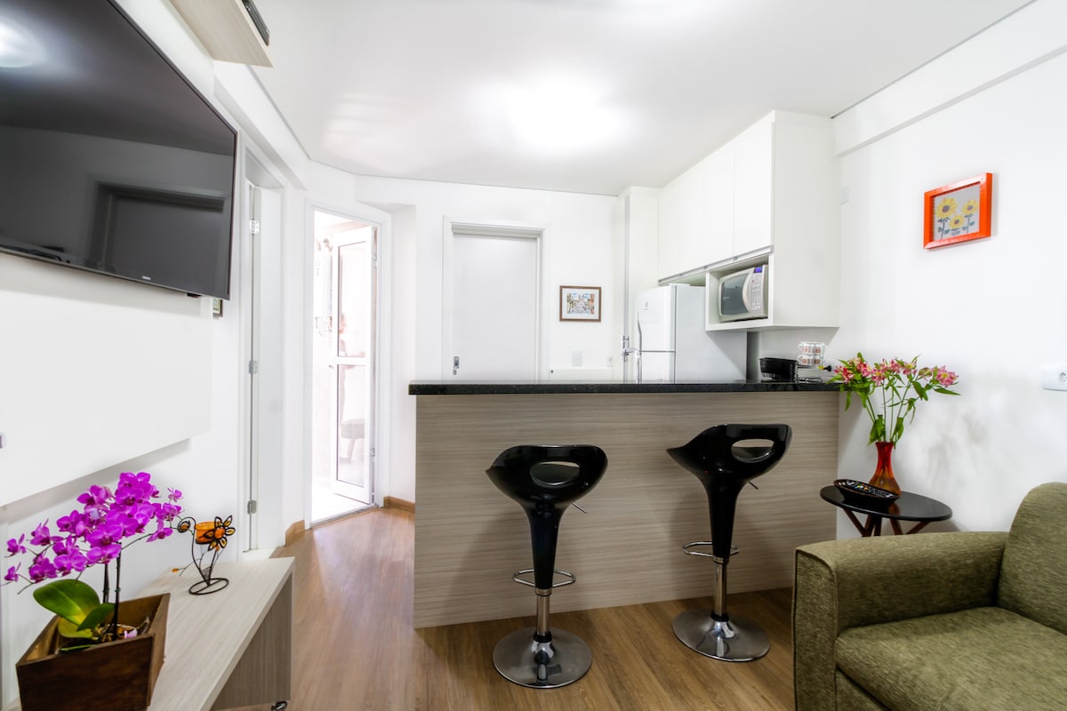 A modern kitchen area features a sleek black countertop with two high-backed bar stools. The setup includes a microwave and other appliances within light-colored cabinetry. A flat-screen television is mounted on the wall, complemented by plants in decorative pots on the counter.