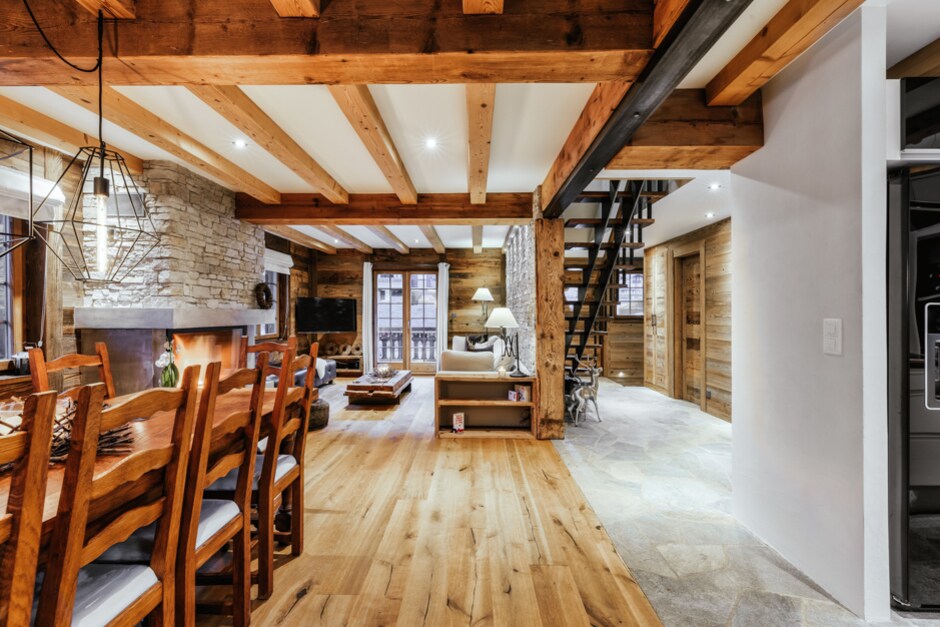 A spacious living area showcases a large wooden dining table, a cozy seating arrangement near a stone fireplace, and exposed wooden beams across the ceiling. Natural light filters in through glass doors, leading to an outdoor area. A staircase with a metal railing adds modern contrast.