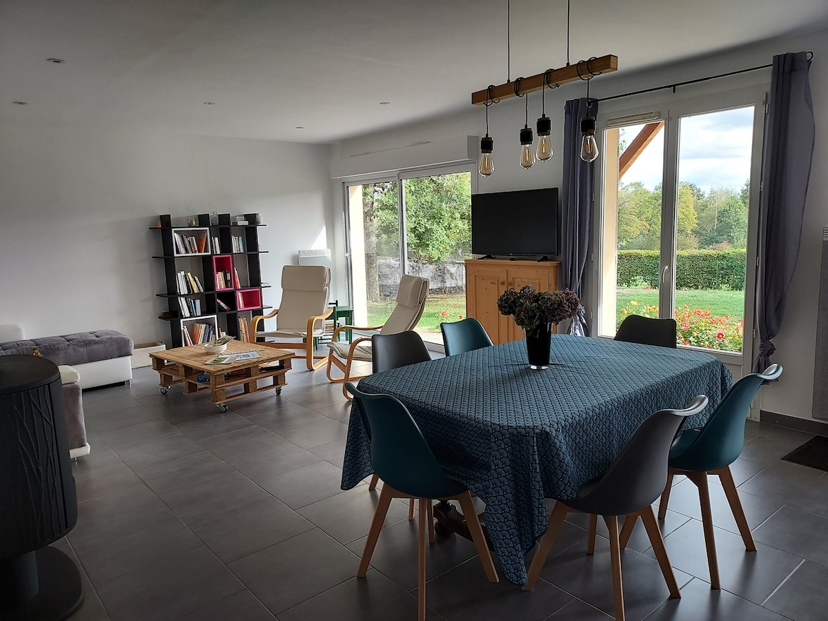 A spacious living area features a dining table surrounded by modern chairs, with a cozy seating arrangement nearby. Large glass doors provide a view of the outdoor greenery, while shelves display books. A television stands against the wall, and overhead lights add brightness.