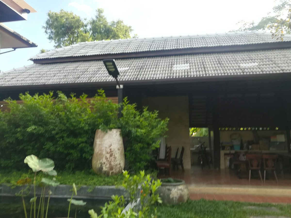 A rustic house blends with its natural surroundings, featuring a tiled roof and surrounded by lush greenery. An outdoor sitting area is visible, along with a stone planter and a glimpse of a well-maintained garden, enhancing the tranquil setting.