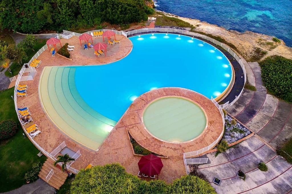 An expansive swimming pool features a unique shape, with clear blue waters surrounded by landscaped areas. Several lounge chairs and umbrellas are positioned around the pool, along with illuminated pathways leading to the adjacent rocky shoreline.