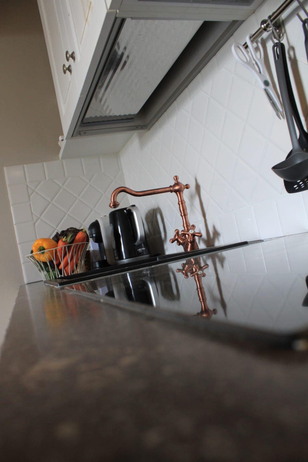 A well-equipped kitchen features a polished countertop with a reflective surface. A black kettle and a range of cooking utensils are neatly arranged, while a bowl of fresh fruits adds a touch of color. Subtle lighting highlights the modern fixtures and tiled backsplash.