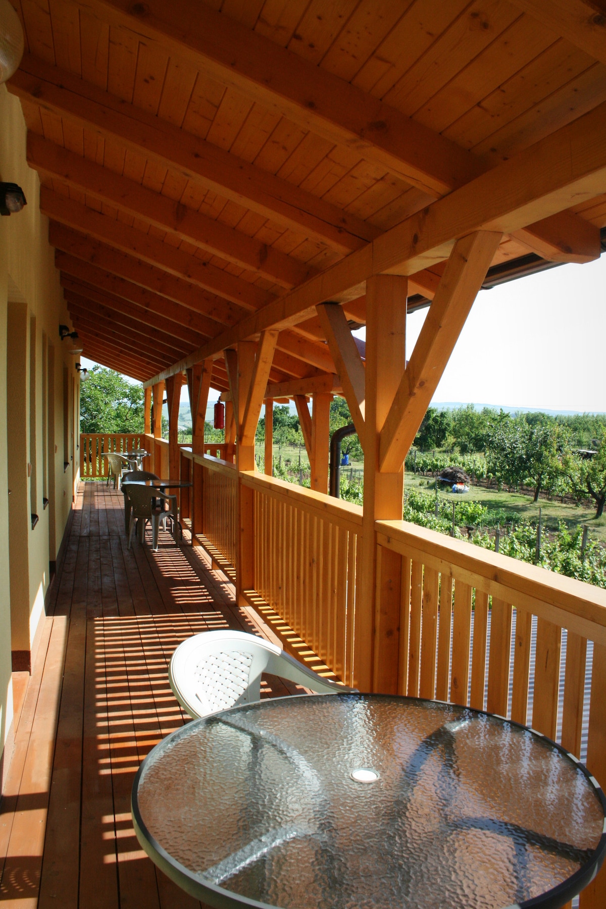 A covered balcony features wooden beams and a railing, providing a scenic view of surrounding greenery. A glass table with chairs invites relaxation in the outdoors, with sunlight casting shadows on the wooden floor.