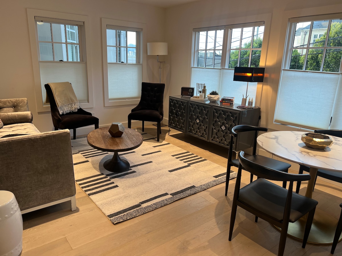 The living and dining area features a blend of modern furnishings, including two black upholstered chairs and a circular coffee table at the center. Natural light fills the space through multiple windows with light-filtering shades, while a patterned rug adds visual interest.