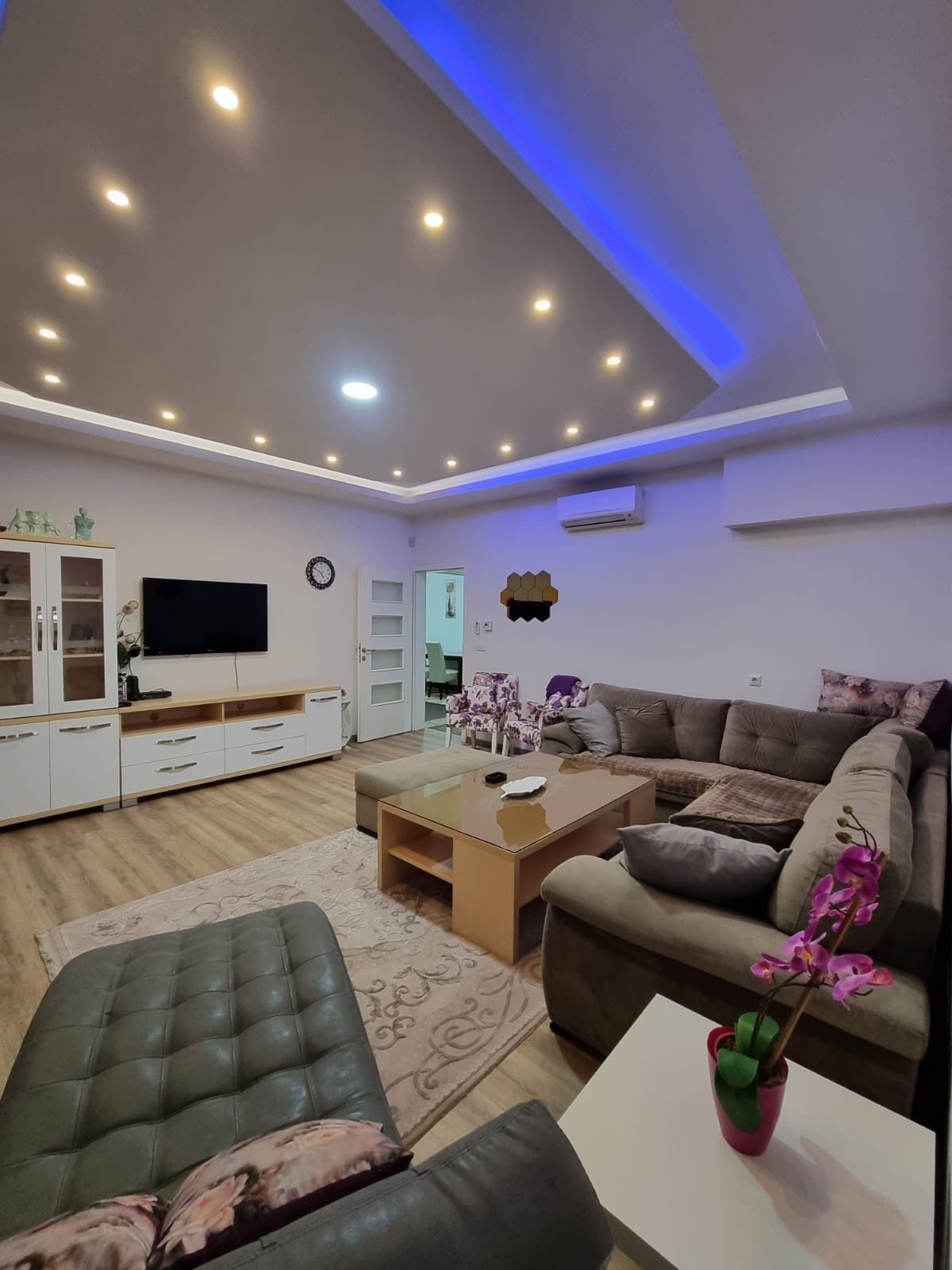 A modern living room features a spacious sectional sofa with soft cushions, a coffee table, and a side chair. A large flat-screen TV is mounted on the wall, and soft blue accent lighting enhances the ceiling design. Floral decor adds a touch of warmth.