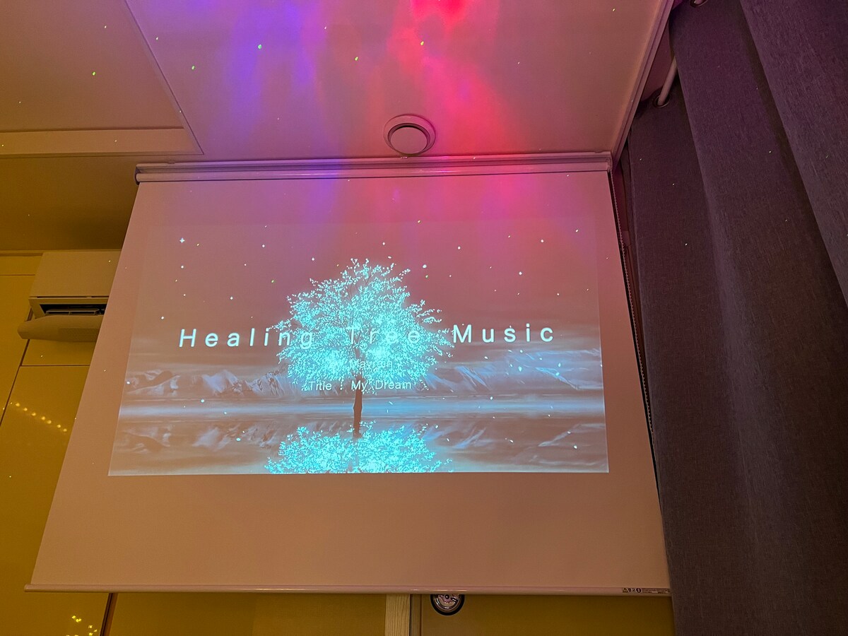 A wall-mounted projector screen displays a tranquil image of a tree against a serene backdrop. Colorful lights shimmer above, creating a calming atmosphere for relaxation or entertainment.