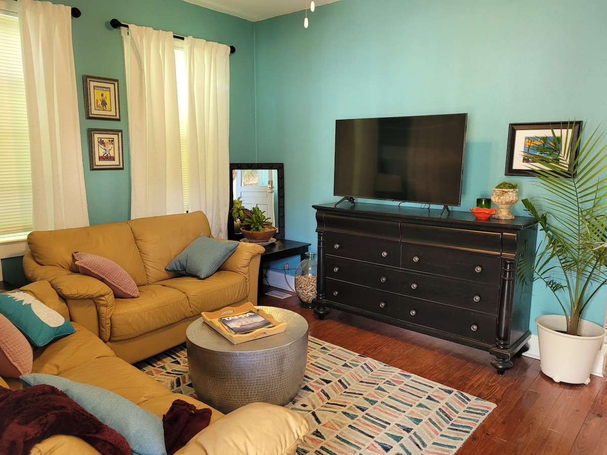 A cozy living area features a light brown leather sectional sofa adorned with colorful pillows. A round ottoman sits at the center, accompanied by a decorative mirror and a large television on a dark wooden dresser. Sunlight filters through sheer curtains, illuminating the space.