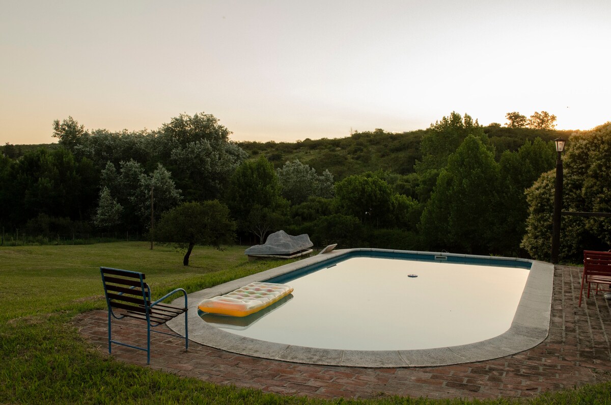 A calm outdoor area features a swimming pool surrounded by lush greenery and trees. An inflatable mattress floats on the water, while a few patio chairs and a table are set nearby, creating a space for relaxation and enjoyment.