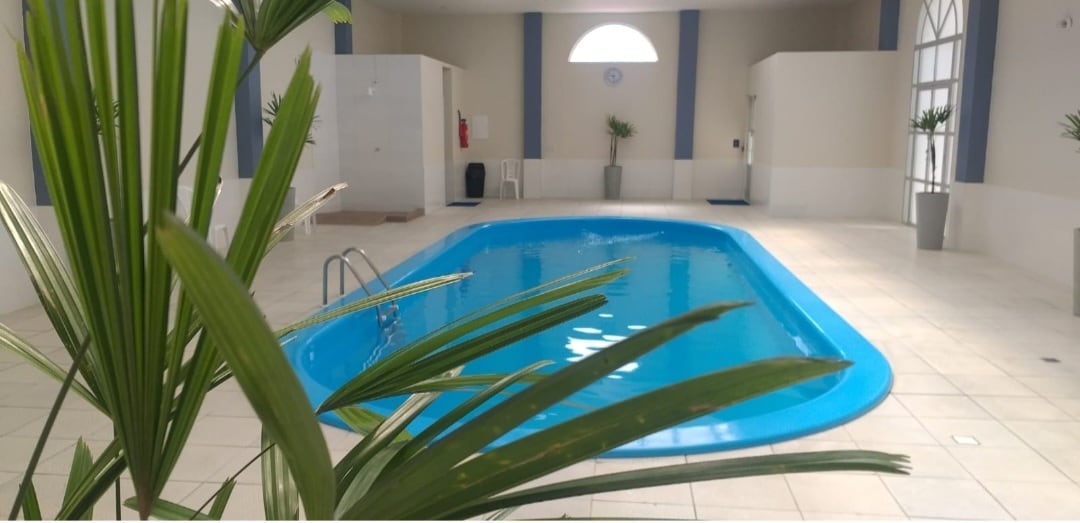 An indoor swimming pool occupies the center of the space, featuring clear blue water. Surrounding the pool, potted plants add a touch of greenery. Natural light is filtered through a large, arched window, illuminating the tile floor.