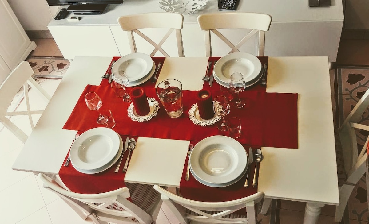 A dining table is elegantly set with white plates, glasses, and utensils, arranged on a red table runner. Centerpieces include candles and a clear vase filled with water. The white chairs complement the light decor of the room, creating a welcoming dining atmosphere.