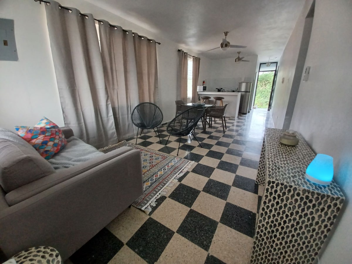 A living area features checkered tile flooring, complemented by a neutral-toned sofa and decorative cushions. A dining table with chairs is positioned nearby, and natural light filters through window curtains, highlighting a serene atmosphere. The open layout connects to the kitchen area in the background.