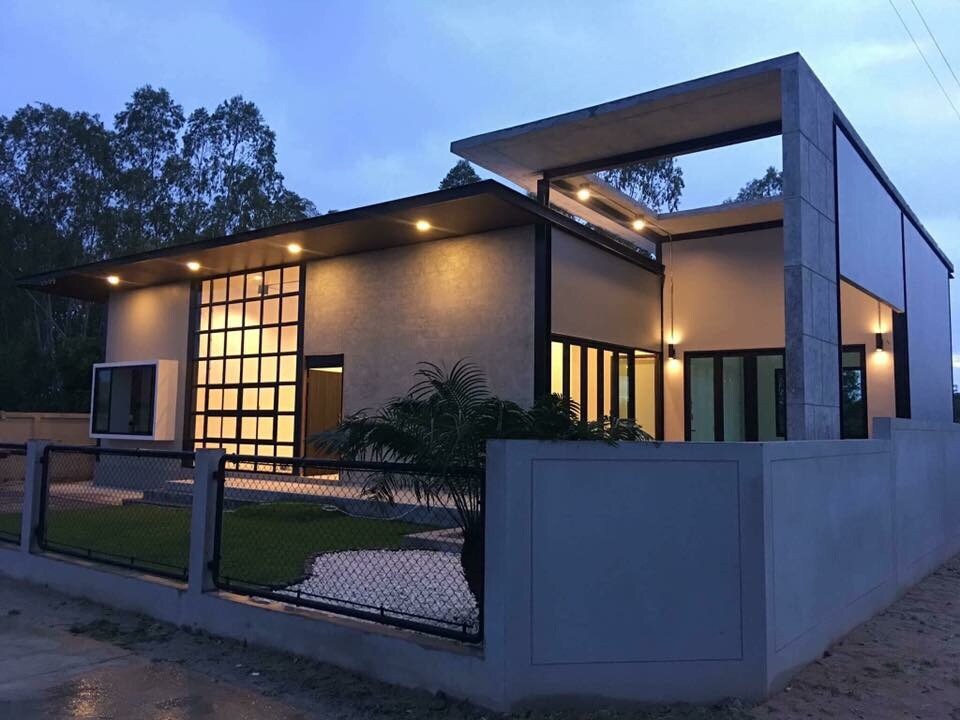 The modern house features a blend of concrete and glass elements, with a large grid window highlighting the entrance. Soft lighting enhances the exterior, and a landscaped area is visible in the foreground.