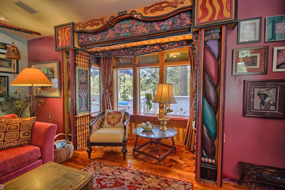 A vibrant seating area features intricate wall details and a large, round wooden table. Bright light filters through the multiple windows, illuminating the rich colors of the walls and decorative elements. A comfortable chair is paired with a patterned sofa, creating a cozy atmosphere.