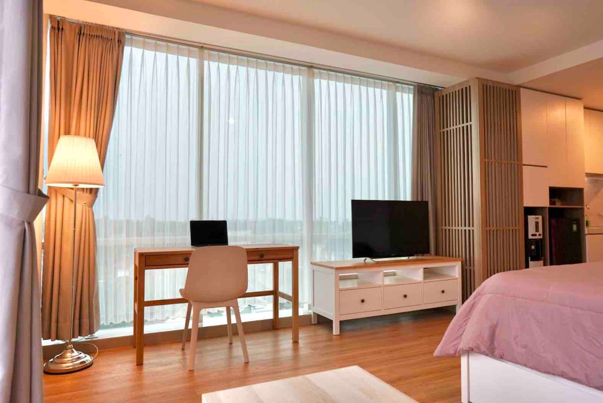 An inviting room features a large window with sheer drapes allowing natural light to fill the space. A wooden desk with a chair is positioned next to a television on a white media console, enhancing the comfortable atmosphere. Warm wooden flooring complements the decor.