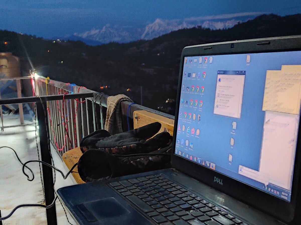 A laptop is placed on a wooden table, capturing a workspace with a scenic view of the snow-capped mountains in the distance. The twilight sky casts a soft hue, enhancing the serene environment for remote work.