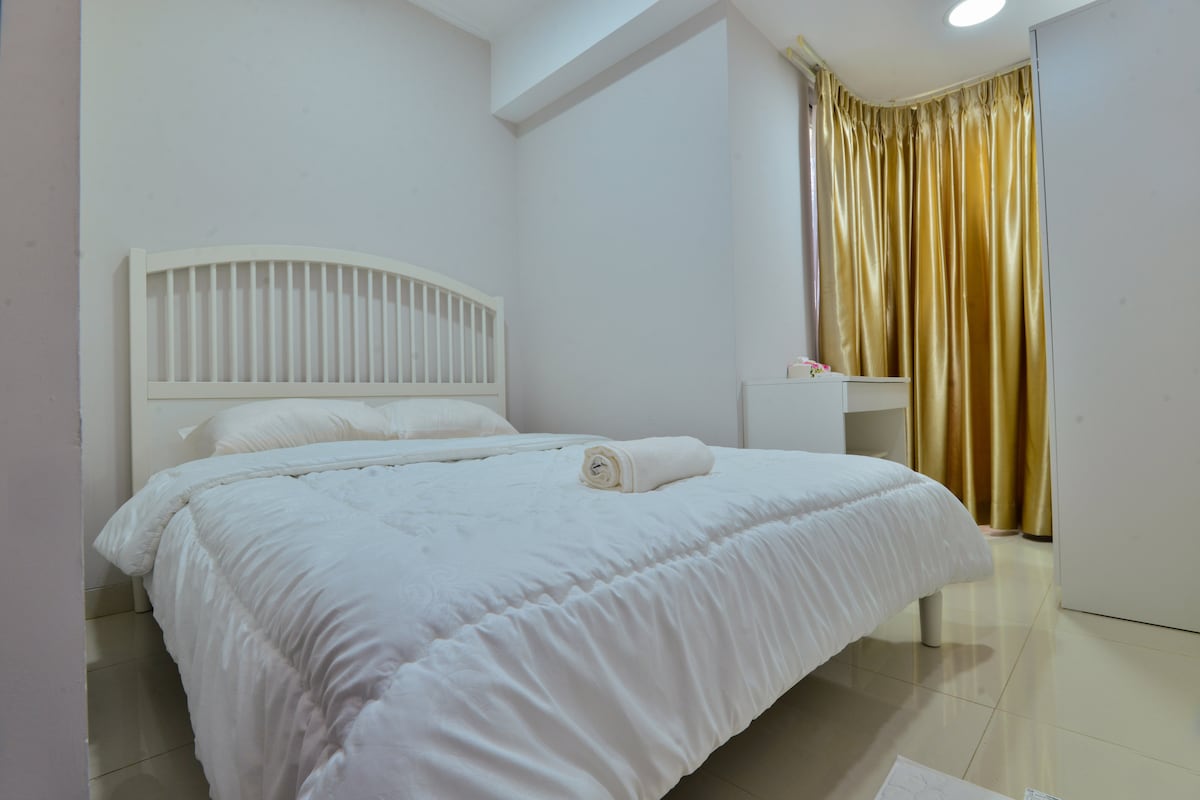 A well-lit bedroom features a spacious bed covered with a white comforter, accompanied by a neatly rolled towel. Natural light enters through golden curtains, creating a bright atmosphere. A small desk is positioned against the wall, complementing the clean design of the room.