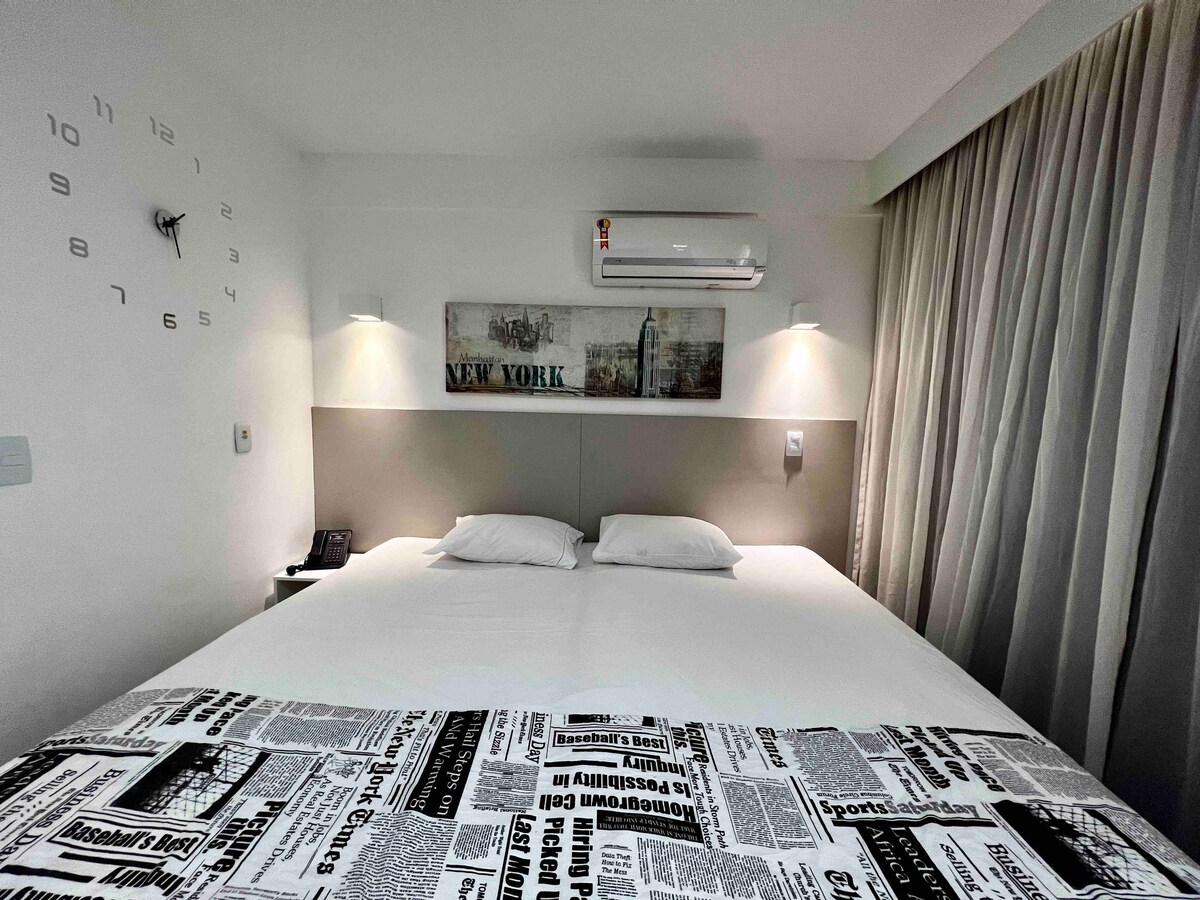 A spacious sleeping area features a large, neutral-toned bed with two pillows. A decorative headboard displays an image of New York, complemented by subtle wall art. Soft curtains frame a window, providing natural light, while a clock is visible on the wall.