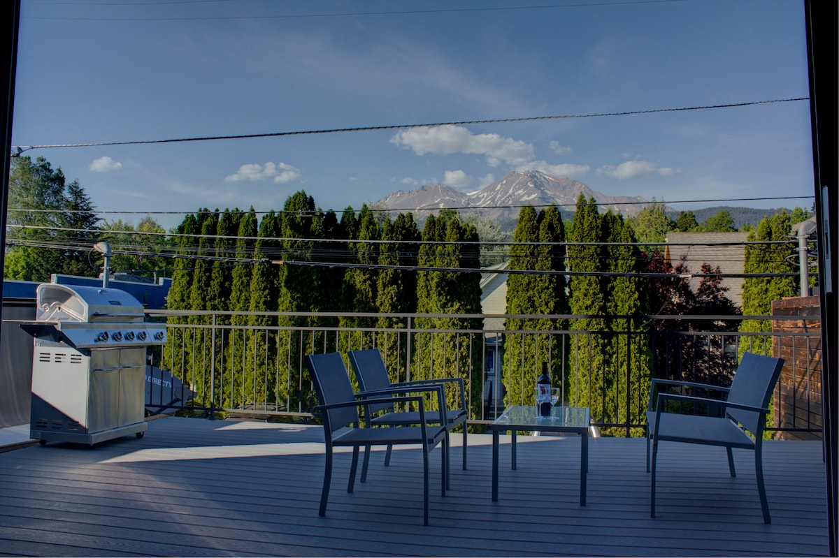 A spacious deck is shown with four chairs arranged around a table, offering views of Mt. Shasta in the background. A barbecue grill is positioned to the left, while tall green trees provide a natural backdrop.