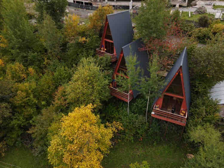 Two A-frame cabins are nestled among vibrant trees, showcasing a blend of greenery and autumnal colors. Each cabin features a balcony with minimalistic wooden railings, enhancing the connection with the surrounding natural environment.