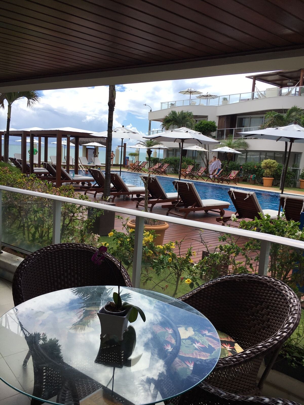 A serene pool area is framed by lounge chairs and shaded umbrellas. The inviting turquoise water of the pool reflects the surrounding greenery and flowers. A terrace table with a plant centerpiece provides a cozy spot to relax, showcasing the peaceful outdoor environment.