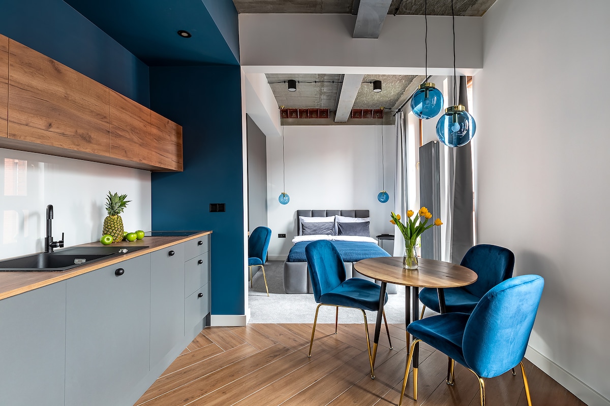 A modern kitchen area features sleek cabinetry with a wooden countertop. A round table with four blue velvet chairs is complemented by a vase of yellow flowers. Light blue pendant lights hang above the table, and a spacious bed is visible in the background.