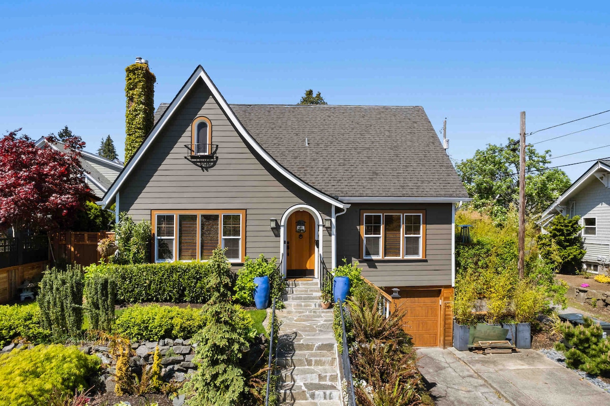 A charming cottage-style home features a sloped roof and large front windows. The front yard is landscaped with a variety of greenery, including colorful plants in blue pots. Stone steps lead up to the inviting entrance, framed by a warm wooden door.