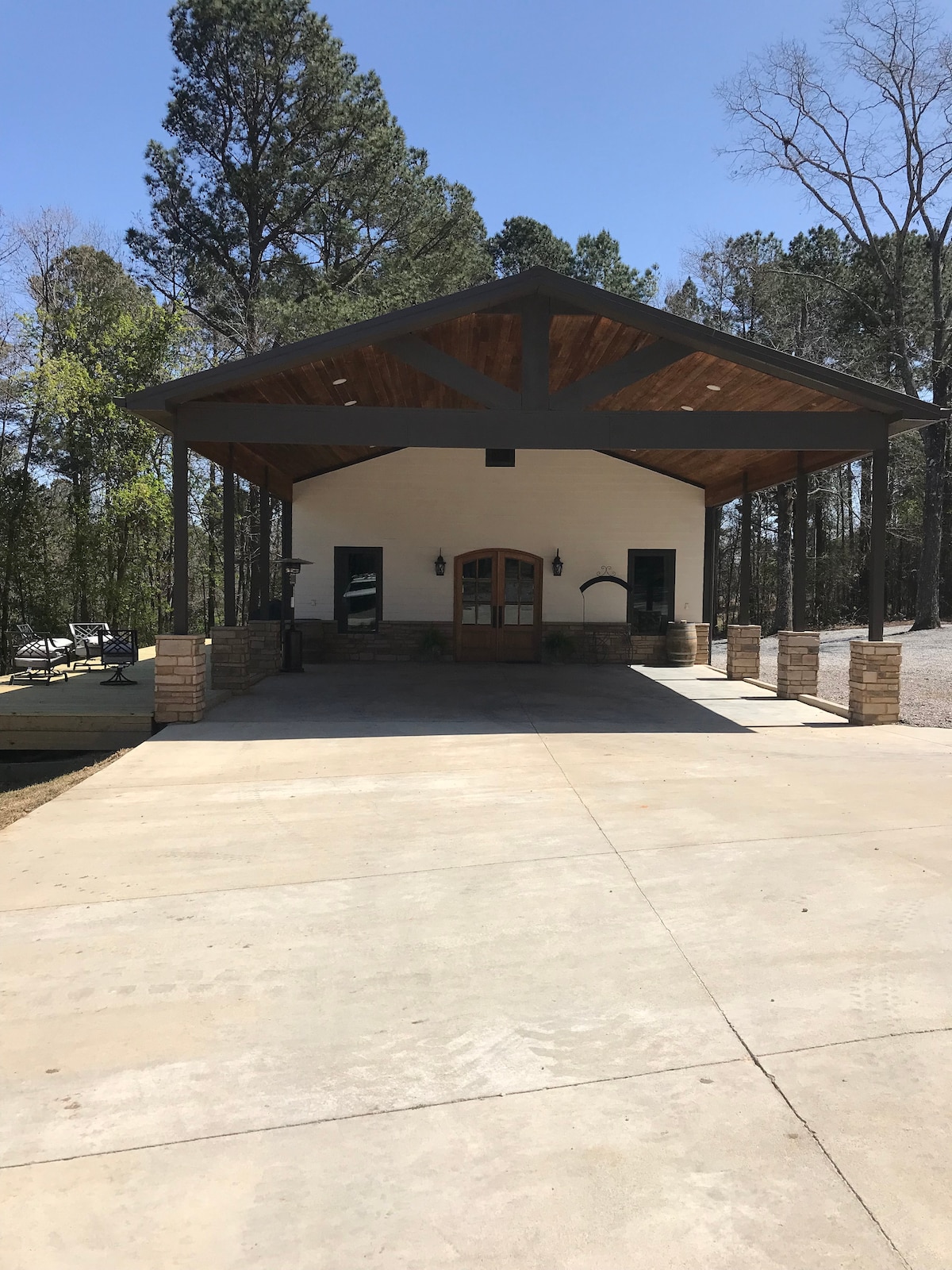 A welcoming entrance to the cottage showcases a wide concrete driveway leading up to a covered porch. The structure features wooden beams and stone accents, framed by tall trees. Enjoy the shaded area with outdoor seating visible to the side.