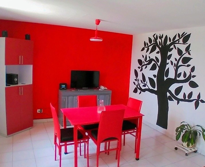 A bright dining area features a red table and matching chairs, creating a bold focal point. A gray cabinet and television are positioned against the wall. An organic tree design is painted on the white wall, with a potted plant placed in the corner, adding a natural element.