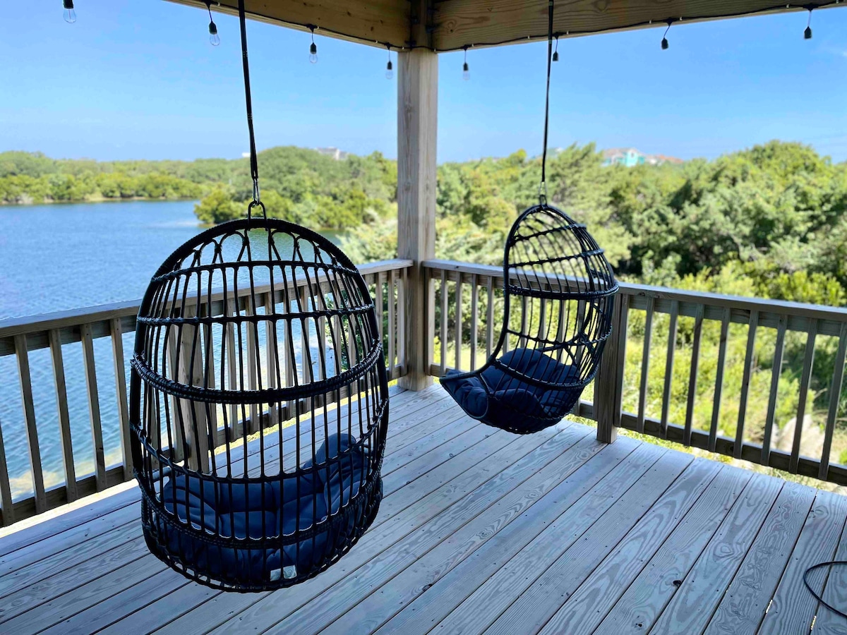 Two hanging chairs are seen suspended from a wooden porch ceiling, overlooking a serene lake surrounded by greenery. The deck features a weathered wooden floor and is bathed in natural light, enhancing the tranquil outdoor setting.