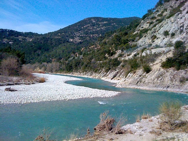A serene river flows through a rocky landscape, surrounded by lush greenery and rolling hills. The clear turquoise water contrasts with the light-colored stones along the riverbank, creating a peaceful natural setting. The distant mountains provide a backdrop to this tranquil scene.