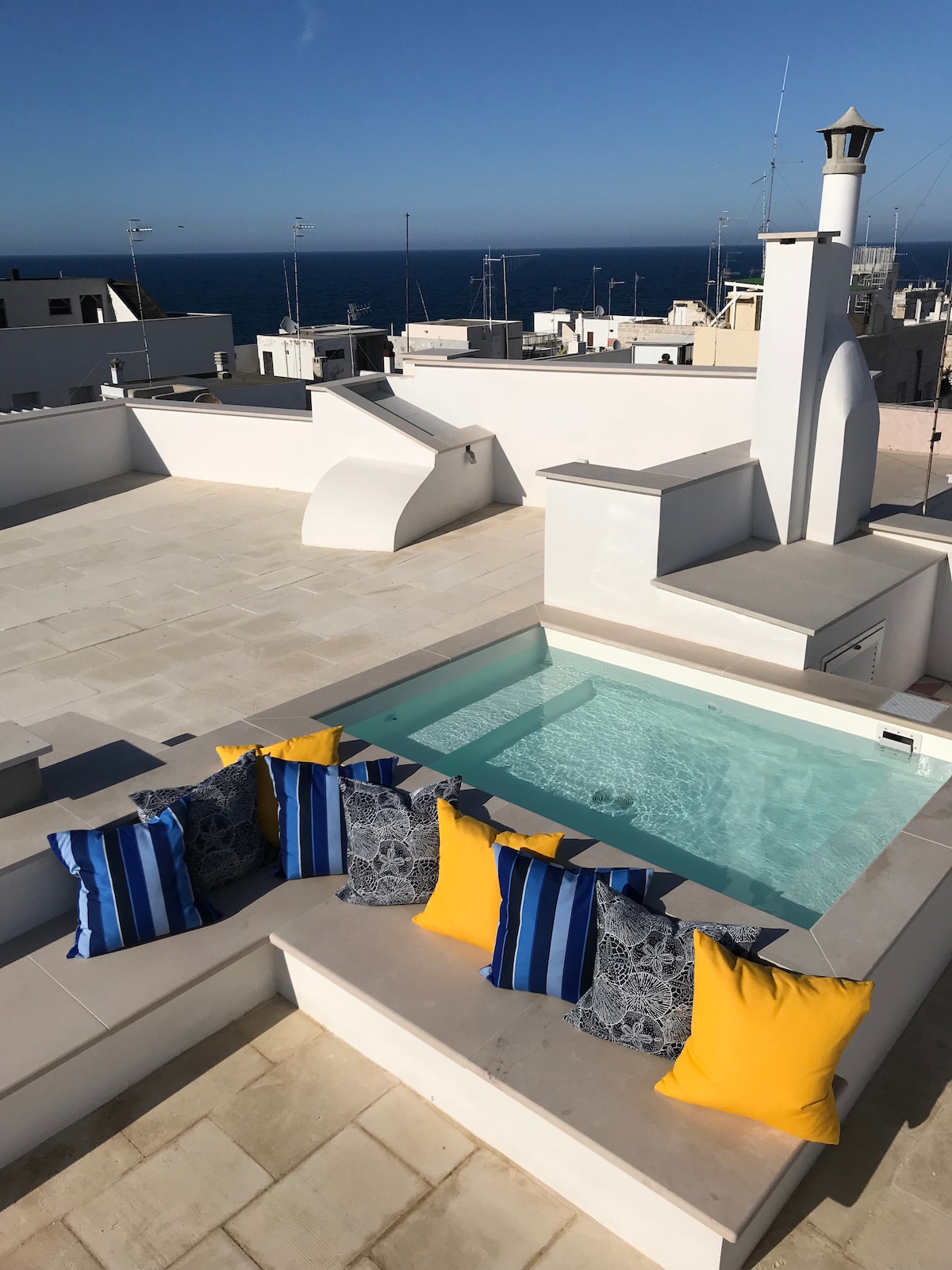 A rooftop terrace features a small plunge pool surrounded by vibrant cushions in blue and yellow. The views extend over whitewashed buildings towards the blue sea, offering a bright and airy setting for relaxation.