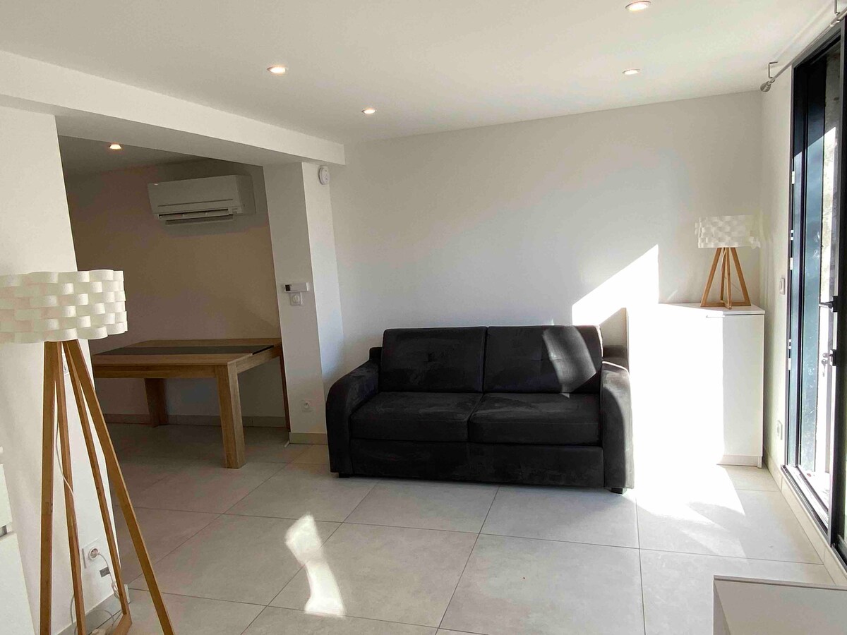 A bright living area is presented, featuring a dark sofa and a wooden table. Natural light fills the space through large sliding glass doors, which lead to the outside. A stylish lamp stands beside the sofa, enhancing the modern design.