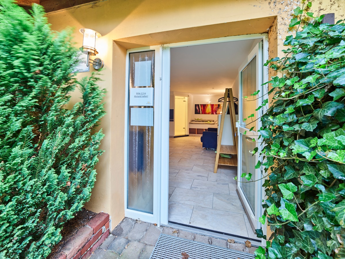 The entrance features double glass doors that open to a bright interior. Lush greenery surrounds the doorway, enhancing the welcoming atmosphere. Inside, natural light illuminates a spacious living area with modern art displayed on the walls.