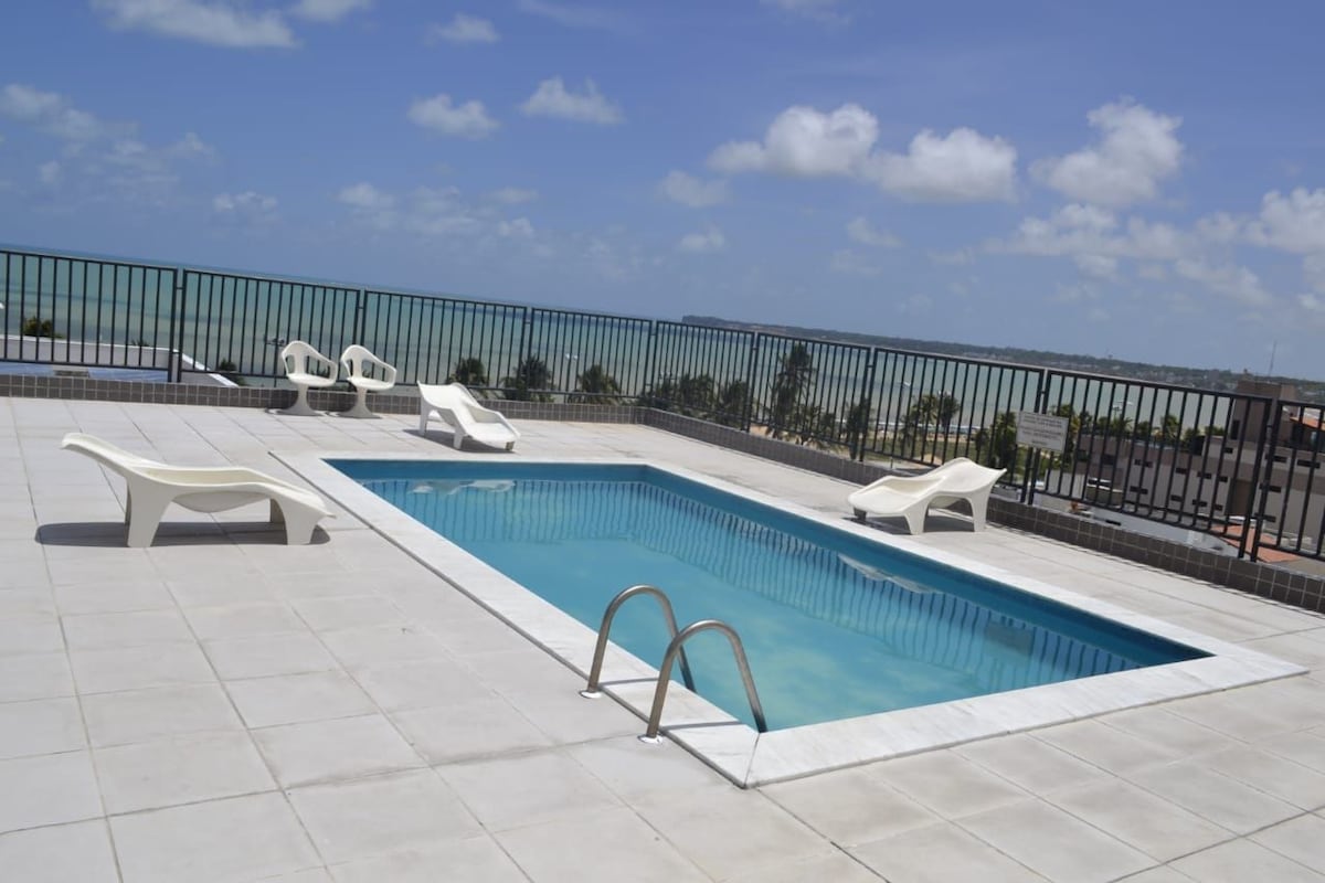 A rooftop pool is framed by a tiled deck, featuring clear blue water and two poolside loungers. The expansive view shows the horizon meeting the sky, with soft clouds dotting the bright blue backdrop.