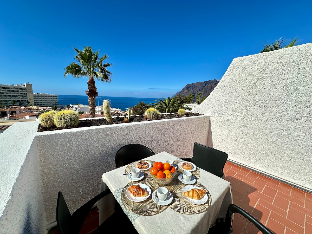 A terrace features a table set for breakfast, displaying pastries and fresh oranges. Two black chairs are positioned around the table. Lush palm trees and cacti are visible, with a panoramic view of the ocean and surrounding landscape in the distance under a clear blue sky.