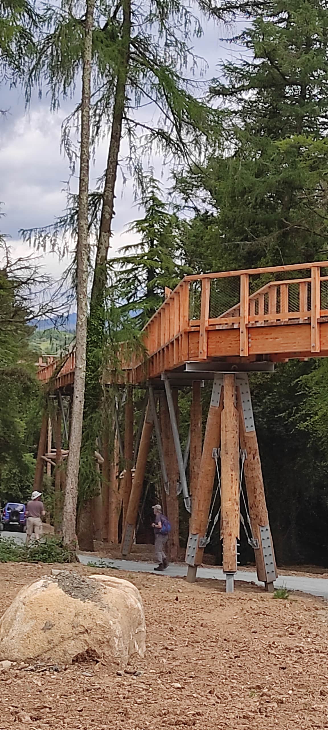 A wooden walkway elevated above the ground is supported by sturdy beams. Lush trees surround the structure, and a pathway leads away from it. Two individuals can be seen strolling along the walkway, while a car is parked nearby, hinting at easy accessibility.