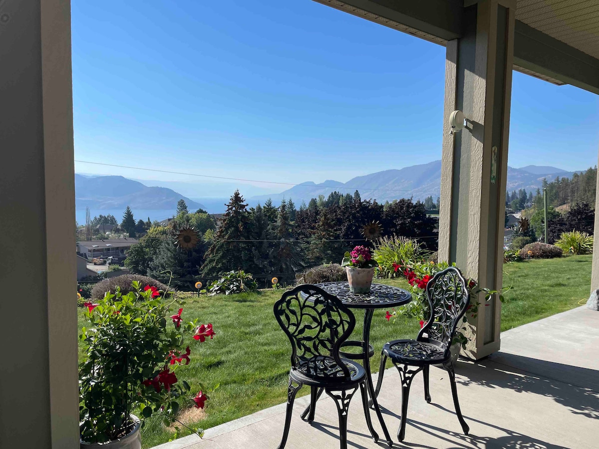 A covered patio features a small round table with two intricate chairs, surrounded by blooming flowers in pots. The expansive view showcases rolling hills and a distant lake under a clear blue sky.