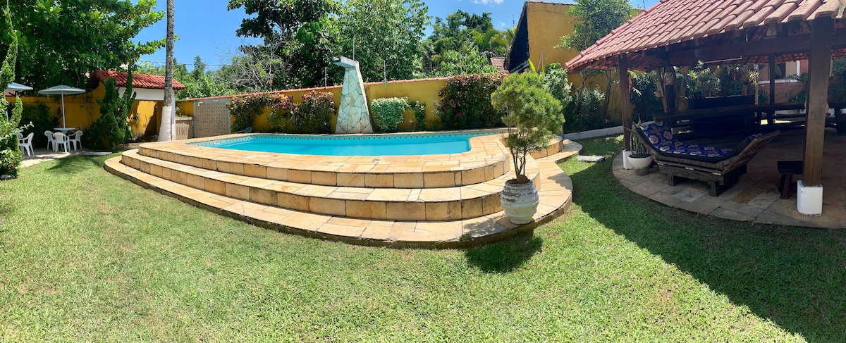 The outdoor area features a spacious lawn surrounding a rectangular pool with stone steps leading up to it. A shaded gazebo with a wooden seating area is visible, along with potted plants and sun loungers nearby, set against vibrant greenery and walls.