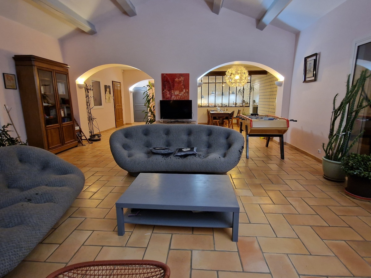 The living area is characterized by two rounded gray sofas and a low coffee table, set on terracotta tiles. A dining space and a well-lit kitchen with a large glass window are visible in the background, enhancing the open layout. Potted plants add greenery to the decor.