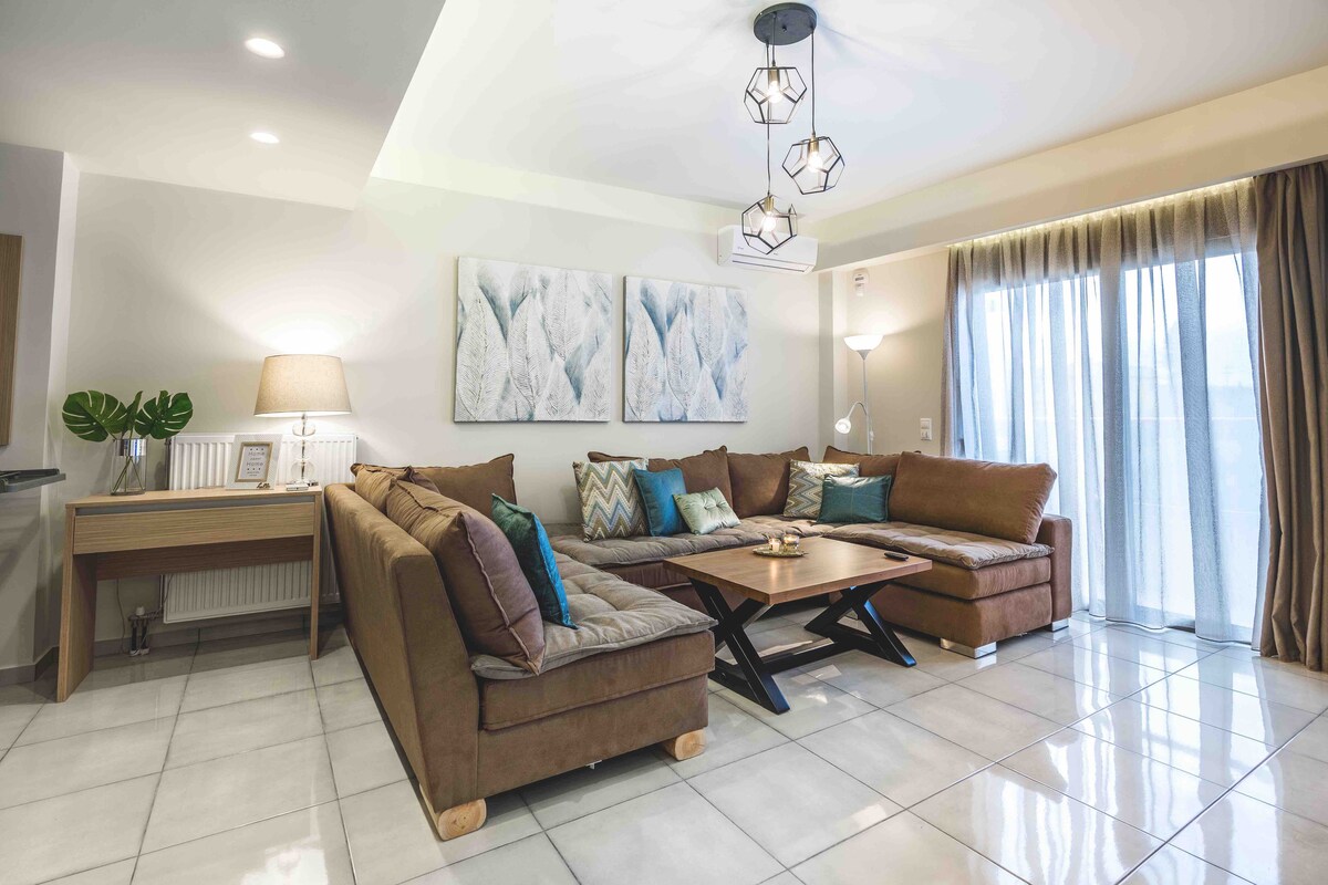 A spacious living area is highlighted by a large, comfortable sectional sofa adorned with decorative cushions. A wooden coffee table is centered on a tiled floor, and soft lighting is provided by modern fixtures. Floor-to-ceiling windows invite natural light, with sheer curtains softly diffusing the view.