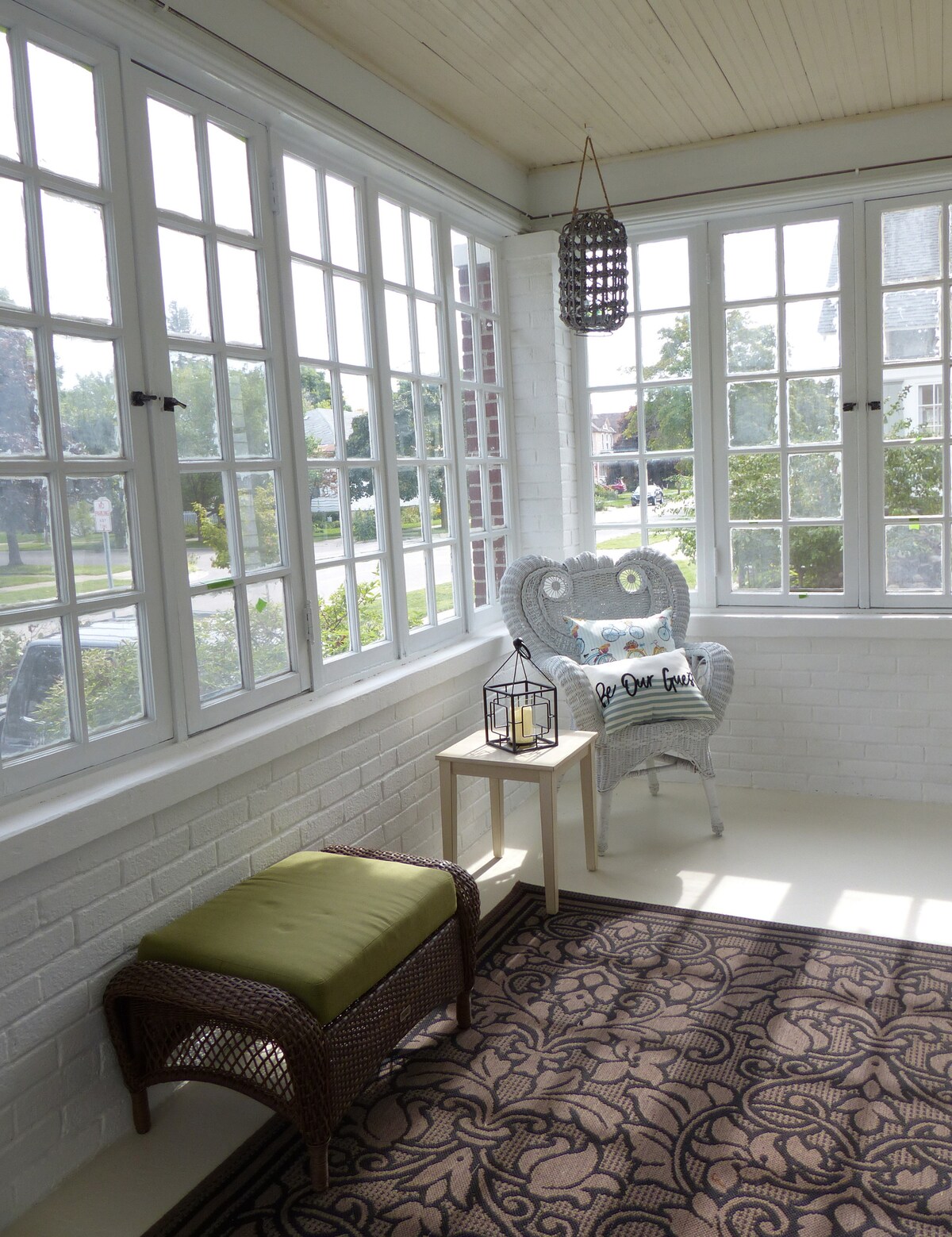 A bright enclosed front porch features large windows allowing ample natural light. A wicker chair and a small table are present, alongside a soft green stool. Decorative elements include a lantern and a patterned rug, creating a serene space for relaxation.
