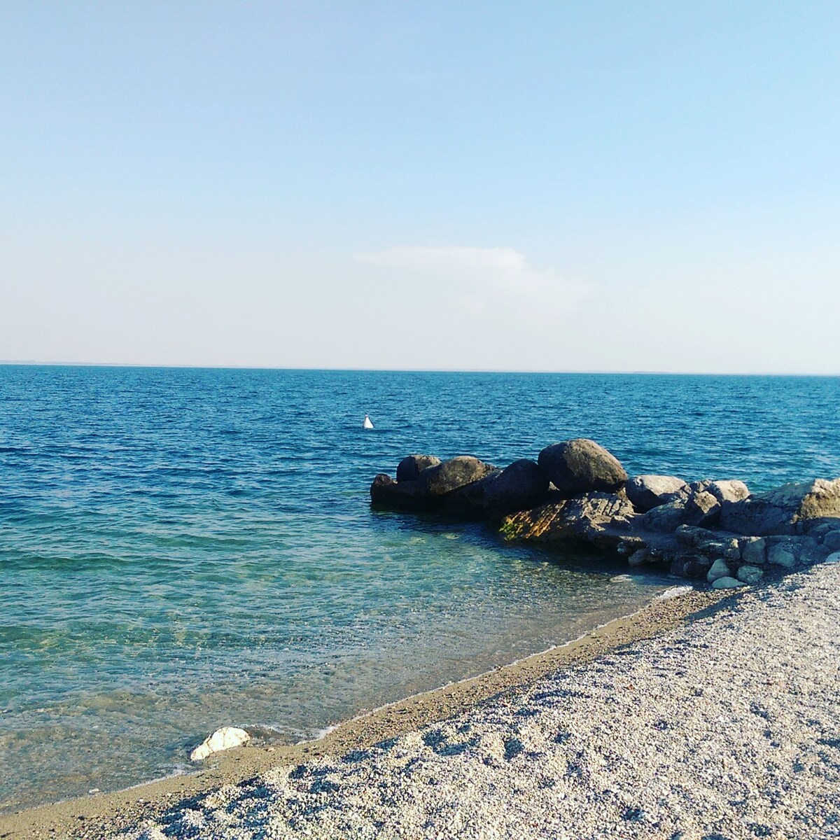 A serene coastal view features gentle waves lapping against a sandy beach. Smooth stones and larger boulders are visible along the shoreline, contrasting with the clear blue water and the sky's soft hues. The scene reflects a peaceful setting ideal for relaxation.