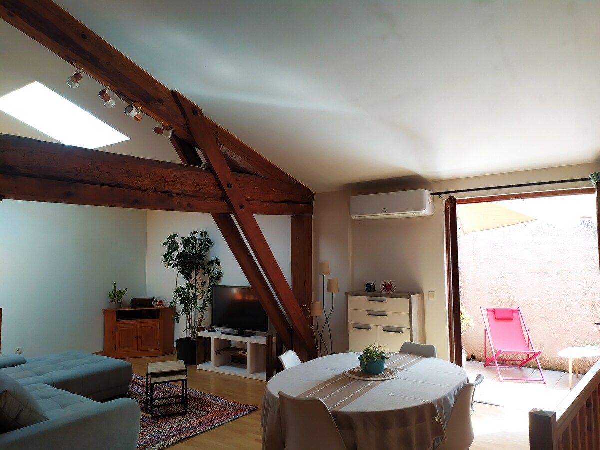 A bright living area features exposed wooden beams and a large skylight, allowing natural light to fill the space. A comfortable seating arrangement includes a sofa and an entertainment unit. A dining table is set for gathering, with a pop of greenery from a nearby plant.