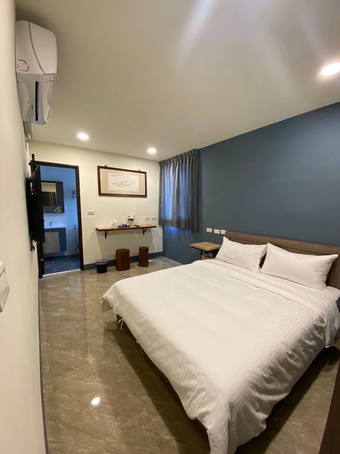 A neatly arranged sleeping area features a spacious bed with white linens and soft pillows. A dark blue accent wall complements the light-colored floor. Natural light enters through a window covered with sheer curtains, while a small table and decorative elements add to the room's functionality.