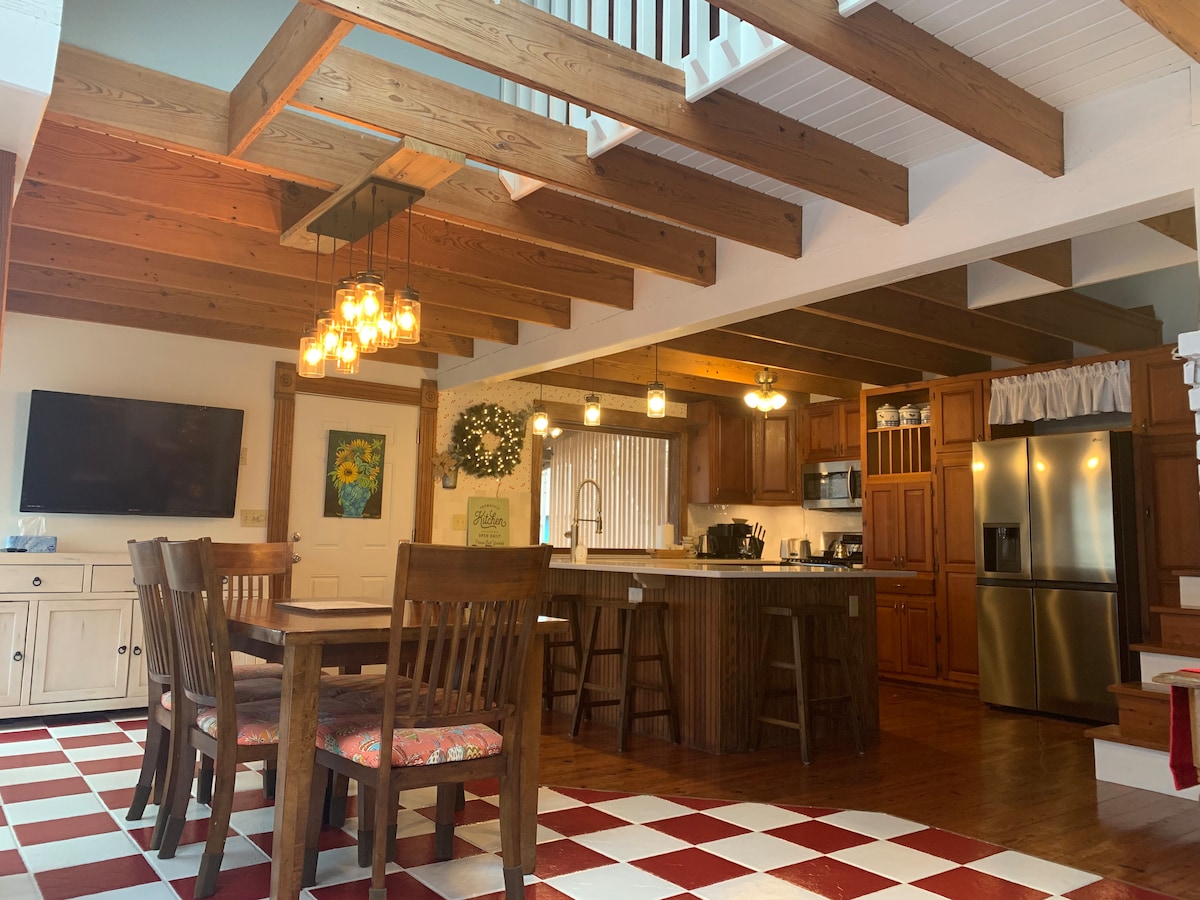 An open concept kitchen and dining area is showcased, featuring wooden beams and a checkered tile floor. A large wooden dining table with high-backed chairs invites gatherings. Bright pendant lighting illuminates the space, while a television is mounted on the wall nearby.