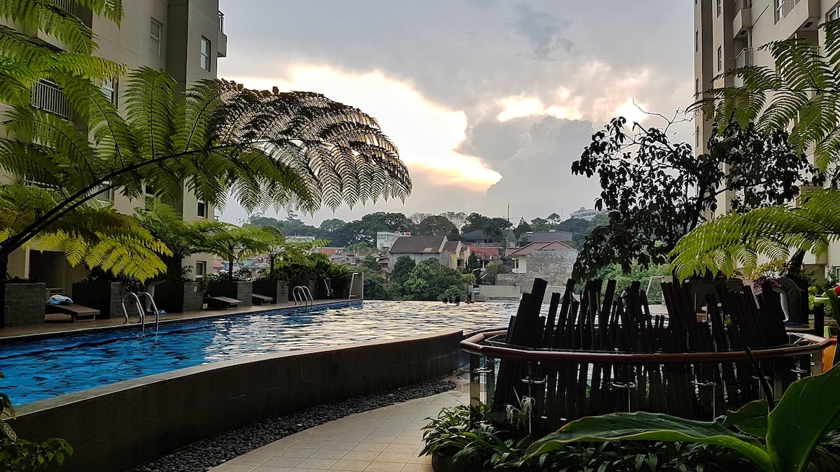 A serene view of the infinity pool is complemented by lush greenery, including ferns and tropical plants. The landscape features rolling hills in the background, with soft clouds over a sunset sky. Comfortable seating arrangements are visible nearby, enhancing the tranquil atmosphere.