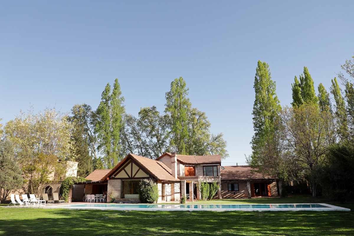 A spacious house featuring a brown tiled roof and wooden accents is surrounded by tall trees and lush green grass. A large swimming pool is positioned in front, reflecting the clear blue sky. The outdoor area is well-maintained, offering an inviting space for relaxation.