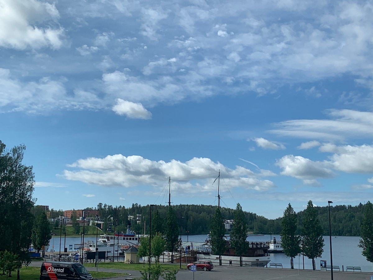 A scenic view of a body of water is framed by lush greenery and gently rolling hills. Several boats are docked along the waterfront, with a distant shoreline featuring buildings. The sky displays a mix of clouds and blue, creating a serene atmosphere.