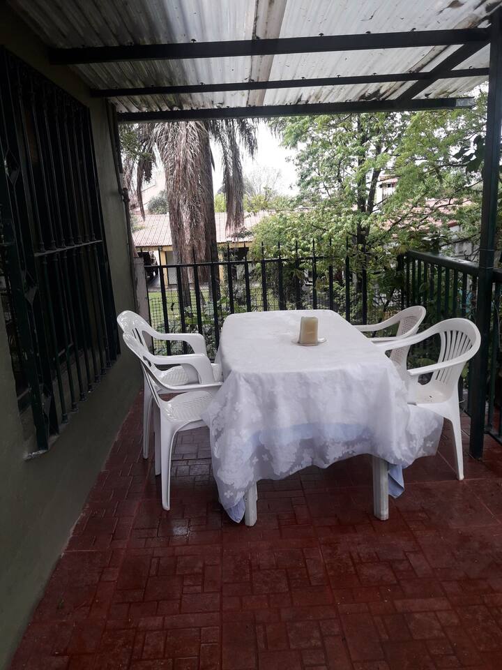 Una vista del mismo balcón con mesa y sillas, rodeado de plantas, y un techo de acrílico que hace posible aún en día lluviosos disfrutar de la vista a las plantas y flores.
